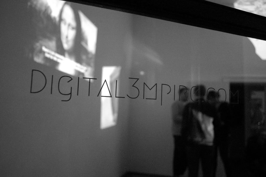 A dark exhibition space, the word digital3mpire projected on the wall